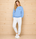 A woman stands against a light wood plank wall wearing a light blue crewneck sweatshirt with small black text on the chest, white jogger sweatpants with matching small text near the cuffs, and white sneakers.