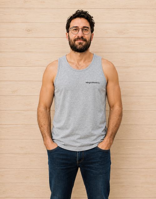 A man with glasses, a beard, and short dark hair stands against a light wood-paneled wall wearing a light gray heathered tank top with 'wing & wood co.' printed in black on the left chest, paired with dark blue jeans.