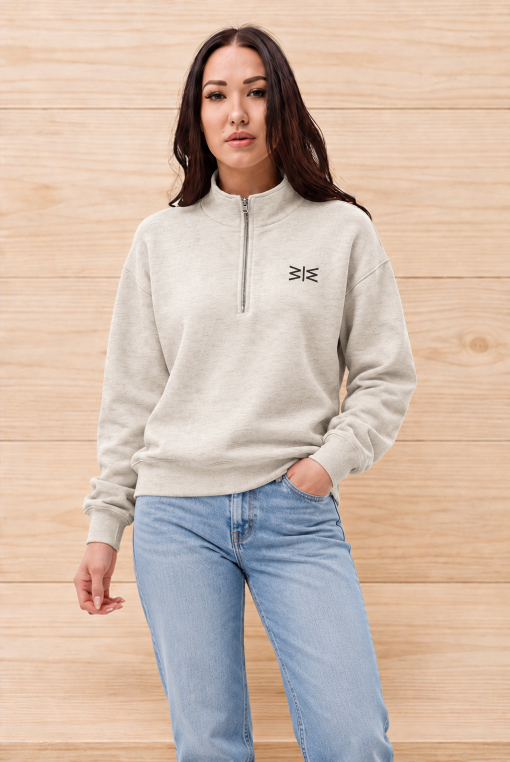 A woman wearing a light beige quarter-zip sweatshirt with a small black embroidered logo on the left chest, paired with light blue jeans, standing against a light wood-paneled background.