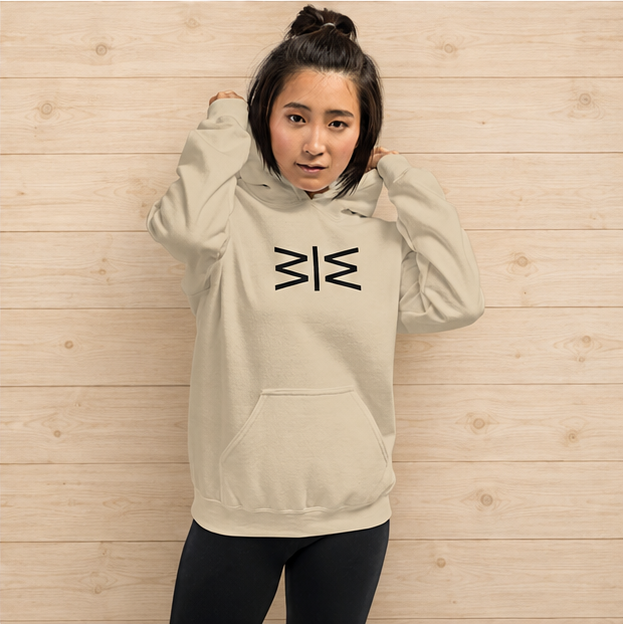 A woman wearing a light beige hoodie with a black geometric logo on the chest, posing against a light wood-paneled wall.