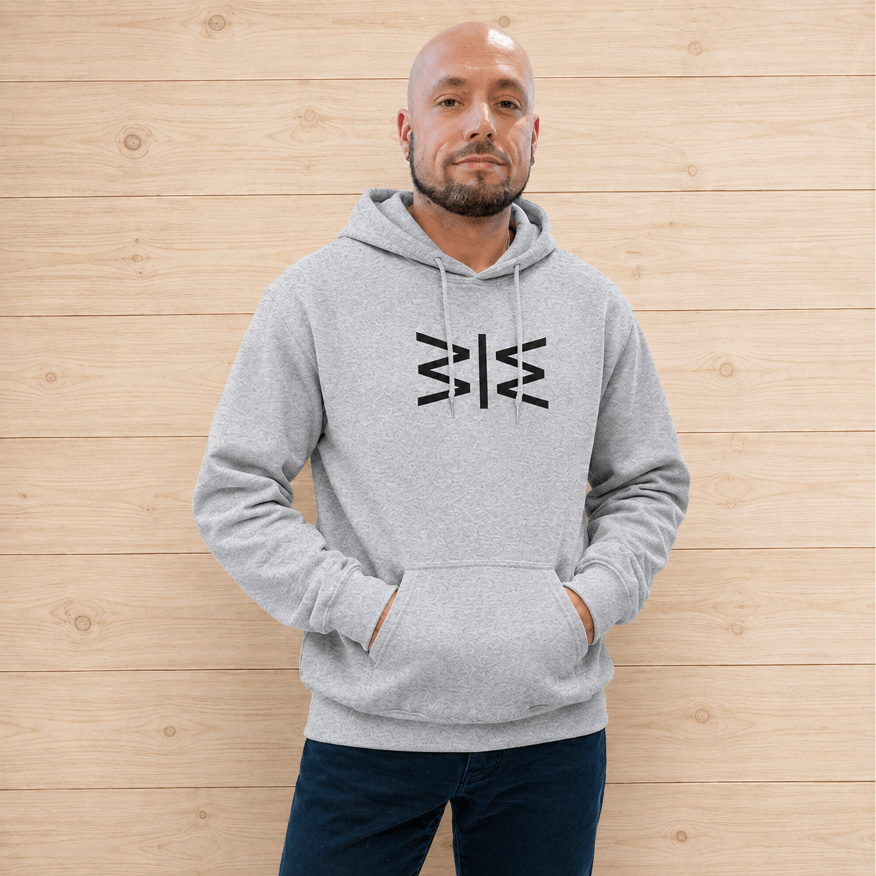 A man wearing a light gray heather hoodie with a black geometric logo on the chest, standing against a light wood plank wall.