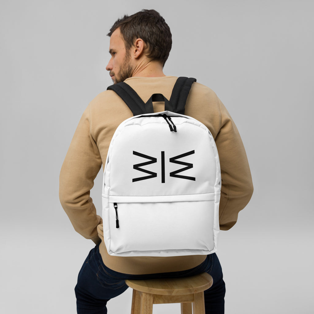 A man wearing a beige sweatshirt and dark jeans sits on a wooden stool, viewed from behind, wearing a white backpack with a black geometric logo on the back.