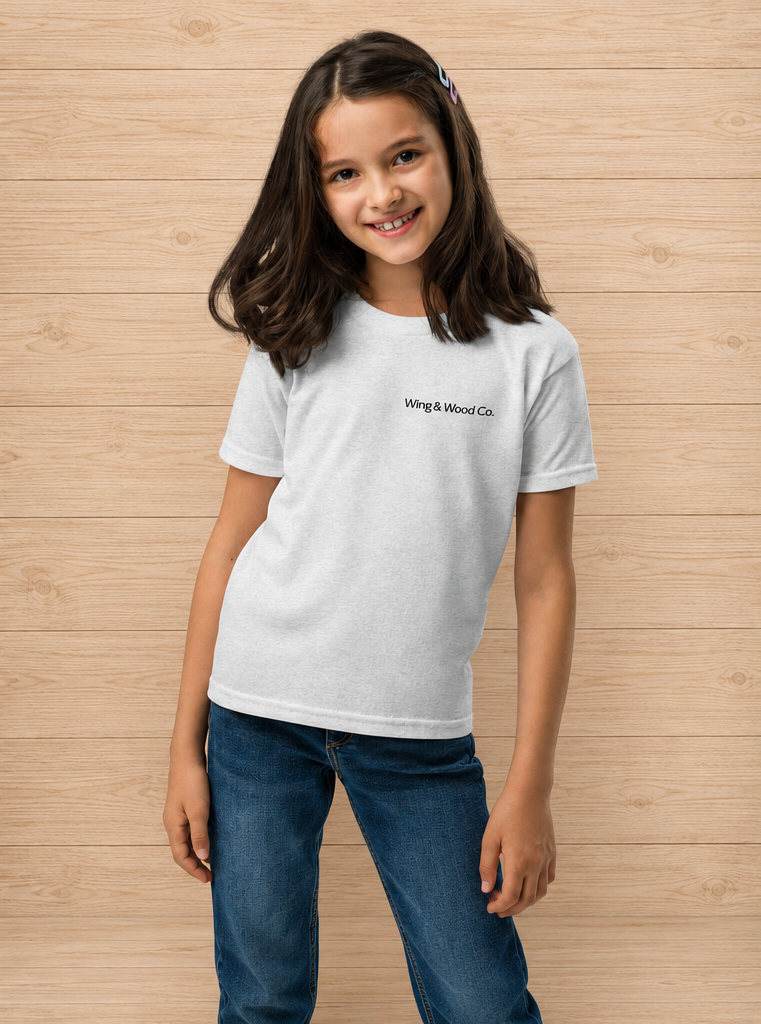 A young girl with long dark hair stands against a light wood-paneled wall wearing a white crewneck t-shirt with 'Whig & Wood Co.' printed in black on the chest, paired with blue jeans.