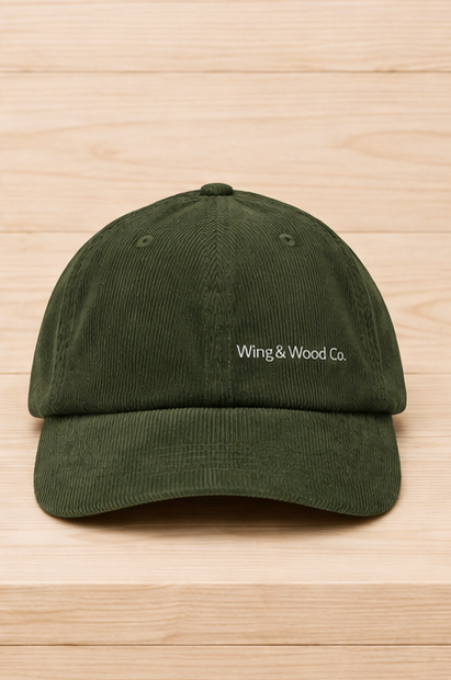 An olive green corduroy baseball cap with 'Wing & Wood Co.' embroidered in white on the front, displayed on a light wood grain background.