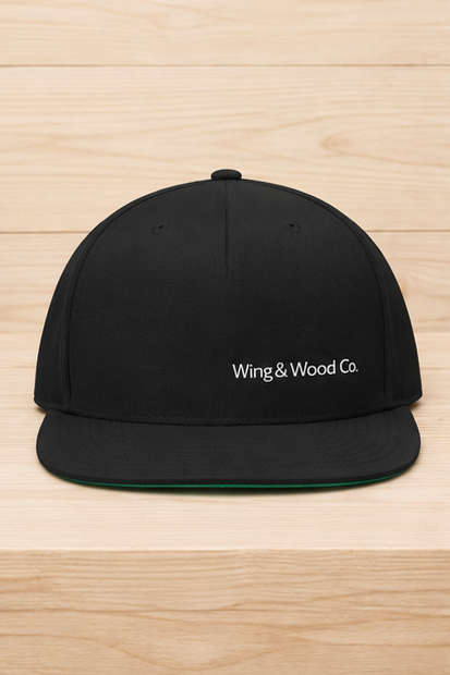 A black flat-brim snapback cap with 'Wing & Wood Co.' embroidered in white on the front, displayed on a light wood plank background.