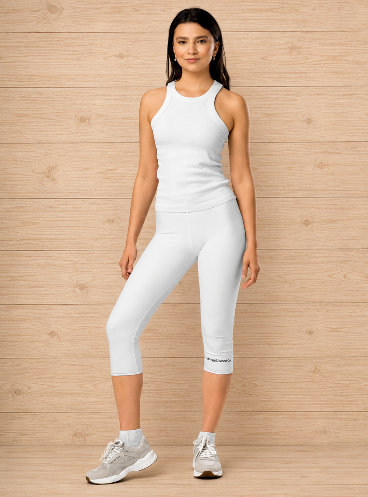 A woman modeling a white ribbed athletic tank top and matching white capri leggings with gray sneakers, standing against a light wood plank wall.