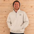 A smiling man wearing a light beige quarter-zip sweatshirt with a small black embroidered logo on the left chest, paired with a dark knit beanie, standing against a light wood plank wall.