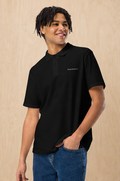 A smiling man with dark curly hair wearing a black short-sleeve polo shirt with 'Design & Wear Co.' printed in white on the left chest, paired with blue jeans, standing against a light wood-paneled background.