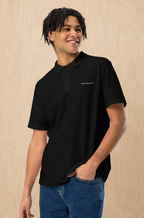 A smiling man with dark curly hair wearing a black short-sleeve polo shirt with 'Design & Wear Co.' printed in white on the left chest, paired with blue jeans, standing against a light wood-paneled background.