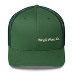 A dark green trucker hat with a structured front panel and curved brim, featuring white embroidered text reading 'Wing & Wood Co.' on the front, set against a plain white background.