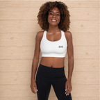 A smiling woman with curly hair stands against a light wood plank wall wearing a white scoop-neck athletic sports bra with a small black logo on the chest, paired with black leggings.