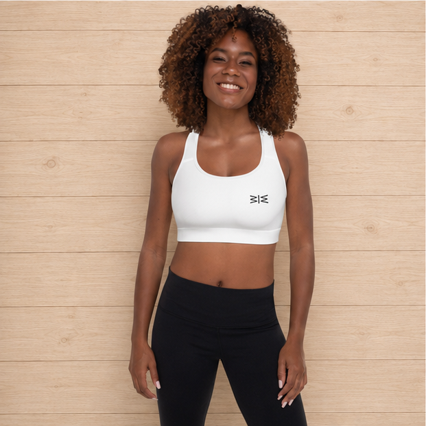 A smiling woman with curly hair stands against a light wood plank wall wearing a white scoop-neck athletic sports bra with a small black logo on the chest, paired with black leggings.