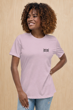 A woman with curly brown hair wearing a light pink crewneck t-shirt with a small black geometric logo on the left chest, paired with light blue jeans, standing against a light wood-paneled wall.