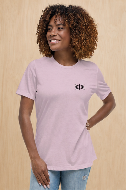 A woman with curly brown hair wearing a light pink crewneck t-shirt with a small black geometric logo on the left chest, paired with light blue jeans, standing against a light wood-paneled wall.