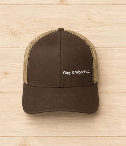 A brown trucker hat with beige mesh back panels and white embroidered text reading 'Wing & Wood Co.' on the front, displayed against a light wood plank background.