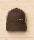 A brown trucker hat with beige mesh back panels and white embroidered text reading 'Wing & Wood Co.' on the front, displayed against a light wood plank background.