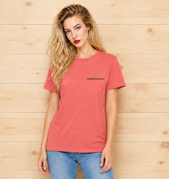 A woman with blonde hair wearing a coral pink t-shirt with 'Wing & Wood Co.' printed in black on the left chest, paired with blue jeans, standing against a light wood plank wall.