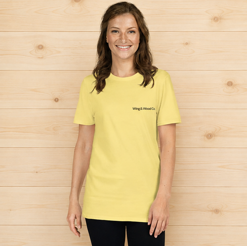 A woman wearing a light yellow short-sleeve t-shirt with 'Wing & Wood Co.' printed in black on the left chest, standing against a light wood plank wall.