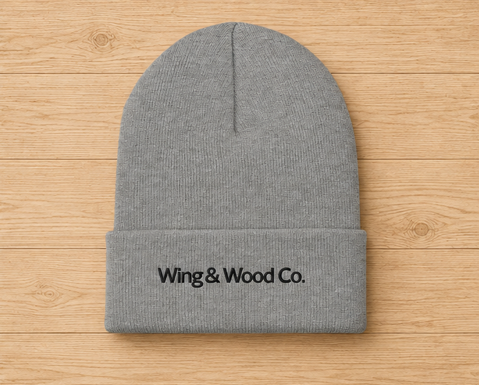 A gray knit beanie with a folded cuff and black embroidered text reading 'Wing & Wood Co.' on the front, laid flat on a light wood plank background.