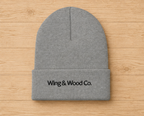 A gray knit beanie with a folded cuff and black embroidered text reading 'Wing & Wood Co.' on the front, laid flat on a light wood plank background.