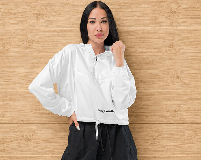 A woman wearing a white zip-up windbreaker jacket with black cargo pants, standing against a light wood plank wall. The jacket features a small black 'wing & wood co.' logo near the bottom hem.